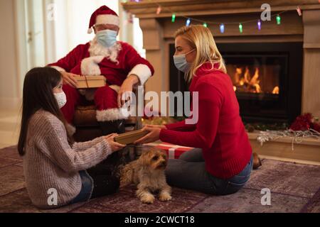 Älterer kaukasischer Mann, seine Erwachsene Tochter und Enkelin zu Hause als Weihnachtsmann gekleidet, trägt Gesichtsmaske, Geschenke zu geben. Soziale Distanzierung Stockfoto