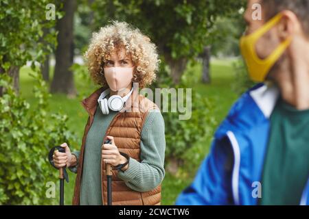 Junge Frau mit lockigen Haaren in Schutzmaske mit Stöcken Während des Sports Spaziergang mit jungen Mann im Freien Stockfoto