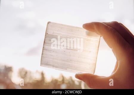 Person hält natürlichen Mineralstein Selenit Platte gegen Sonne und blauen Himmel, Stein hat heilende und reinigende Eigenschaften. Wird auch zum Aufladen verwendet. Stockfoto