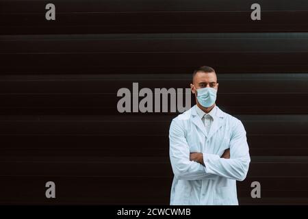 Porträt eines jungen hübschen kaukasischen Arztes in Uniform mit einer Schutzmaske im Gesicht vor einem schwarzen Gebäude. Schutz gegen COVID - 19 Stockfoto