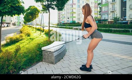 Seitenansicht der jungen muskulösen Frau Training Oberkörper, üben langsam kontrollierte Übungen mit Fitness elastisches Seil Band im Freien. Mädchen genießen die Arbeit mit Widerstand Band mit Bank in der Stadt. Stockfoto