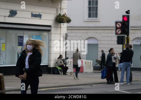 Eine Frau geht entlang der Parade in Leamington mit einer Gesichtsmaske, ihre Haare wehen im Wind. Im Hintergrund spielt ein Busker ein Akkordeon Stockfoto