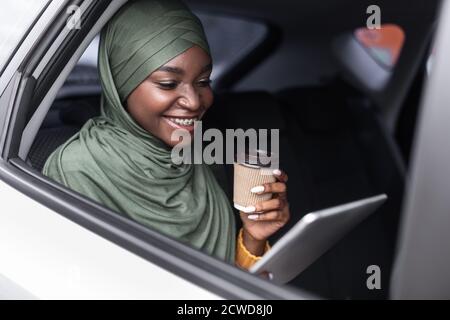 Schwarze muslimische Frau Reiten Auto auf dem Rücksitz mit digitalen Tablet Und Kaffee Stockfoto