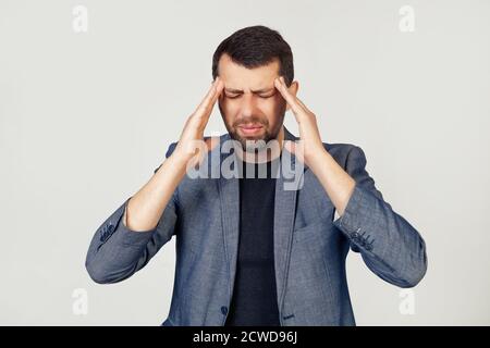 Junger Geschäftsmann mit Bart in einer Jacke. Hände auf dem Kopf, leiden unter Kopfschmerzen in Verzweiflung und unter Stress durch Schmerzen und Migräne. Porträt eines Mannes auf grauem Hintergrund. Stockfoto