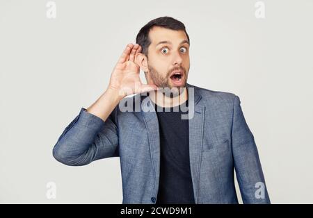 Junger Geschäftsmann mit Bart in einer Jacke, lächelnd mit der Hand über dem Ohr, lauscht Gerüchten oder Klatsch. Taubheitskonzept. Porträt eines Mannes auf grauem Hintergrund. Stockfoto