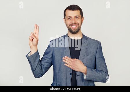 Junger Geschäftsmann mit Bart in einer Jacke, lächelnd, schwört, legt seine Hand auf die Brust und Daumen hoch, schwört einen Treueeid. Porträt eines Mannes auf grauem Hintergrund. Stockfoto