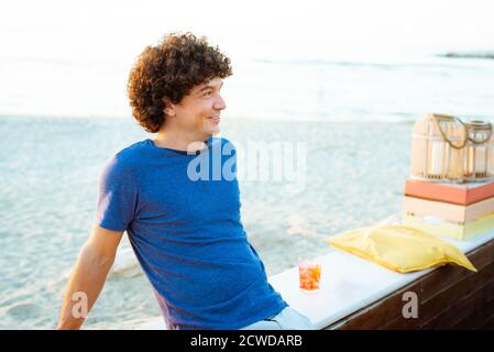 Lockiger kaukasischer Latino-Mann, der bei Sonnenuntergang einen Drink genießt Der Strand lächelnd.Konzept über Freude und Glück zu verführen Cocktail an einem warmen sonnigen Tag Stockfoto