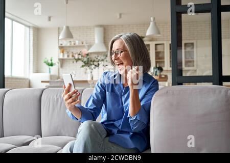 Aufgeregt reifen alten Frau Gewinner Lesung Nachricht auf dem Smartphone zu Hause. Stockfoto