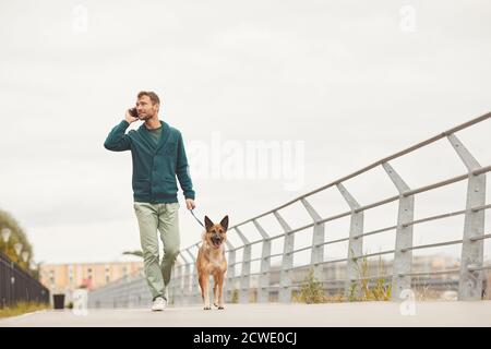Junger Mann, der mit seinem Hund die Straße entlang läuft und Sprechen auf dem Handy Stockfoto