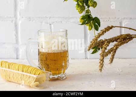 Glas Becher Bier mit Kartoffelchips, Ohren von Gerste und einem Hopfenzweig auf weißem Hintergrund. Stockfoto