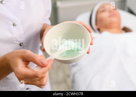 Kosmetikerin Hände halten Schüssel mit Gesichtsmaske Ton mint Farbe Und kosmetische Spatel auf weibliche Client Gesicht in gelten Schönheitssalon Stockfoto