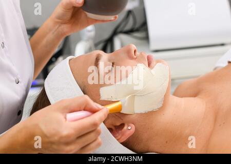 Cosmetologist Hände gelten weiße Reinigung Peeling Alginat Maske auf Client Gesicht mit kosmetischen Pinsel, Seitenprofil Ansicht, Beauty Spa Salon Anti-Aging pro Stockfoto
