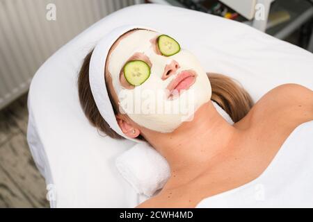 Frau lag auf der Couch unter Handtuch mit weißen Alginat professionelle Peeling Tonmaske und runde Gurkenscheiben decken die Augen, kosmetologische Hautpflege Konzept Stockfoto