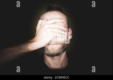 Ein bärtiger junger Mann bedeckt seine Augen mit einer Hand. Stockfoto