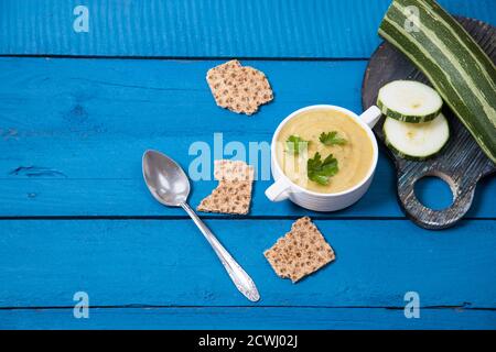 Kürbissuppe-Püree in einem weißen Becher mit Kräutern und Getreidecrackern auf blauem Holzhintergrund, neben einem in Scheiben geschnittenen Kürbis auf einem runden Holzschneidebrettchen Stockfoto