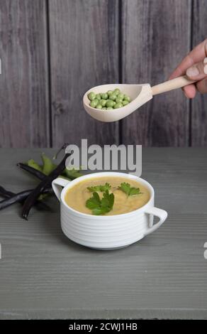 Suppe-pürierte Erbsen und Bohnen in einem weißen Becher auf einem grauen Holzhintergrund, Hand mit einem Löffel mit Erbsen über einer Tasse Suppe . Platz für einen Kopierbereich Stockfoto