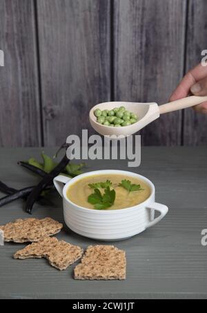 Suppenpürierte Erbsen und Bohnen in einem weißen Becher auf einem grauen Holzhintergrund mit Getreidekräckern, Hand mit einem Löffel mit Erbsen über einer Tasse Suppe . Platz für Stockfoto