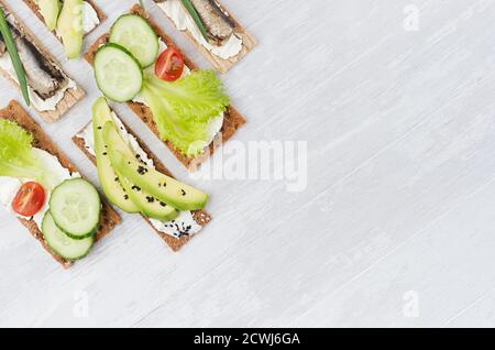 Frische Frühlingsvitamine Sandwiches aus flachem Getreide Roggen trockene Chips Brot mit frischen Scheiben Gemüse, Fischkonserven, Frischkäse auf weißem Holztisch, Stockfoto