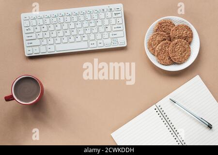Gemütlicher Arbeitsplatz mit Tastatur, Notizblock, Stift, Tasse Kaffee und Keksen auf beigem Hintergrund. Draufsicht, flach liegend, Kopierbereich. Stockfoto
