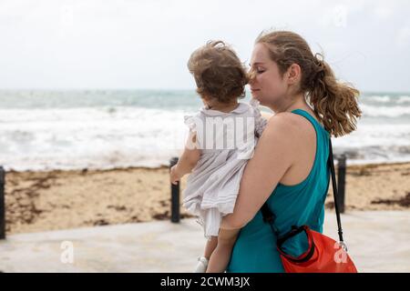 Eine junge Mutter hält liebevoll ein kleines Mädchen in sich Ihre Arme vor dem Hintergrund des Meeres Stockfoto