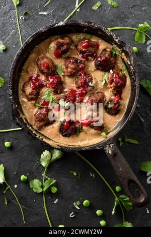 Skandinavisches Komfortessen. Hausgemachte Fleischbällchen in cremiger Soße mit roter Beerenfrucht in Gusseisenpfanne mit rohem Koriander auf schwarzem Hintergrund Draufsicht Cop Stockfoto