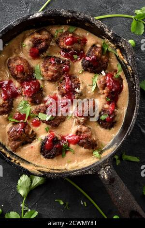 Hausgemachte herzhafte Speisen. Fleischbällchen in Soße mit Preiselbeerensause und Koriander in schwarzer Metallpfanne auf schwarzem Hintergrund Draufsicht Stockfoto