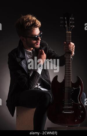 Seitenansicht eines jungen sitzenden Gitarristen, der seine E-Gitarre und sein Halsband hält und auf dunklem Studiohintergrund denkt Stockfoto