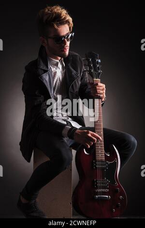 Seitenansicht eines Rock and Roll Gitarristen, der seine E-Gitarre hält und im Sitzen für die Kamera posiert, vor schwarzem Studiohintergrund Stockfoto