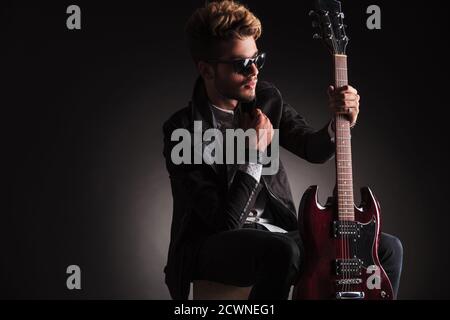 Seitenansicht eines coolen Gitarristen, der seine E-Gitarre hält und auf schwarzem Studiohintergrund von der Kamera wegschaut Stockfoto