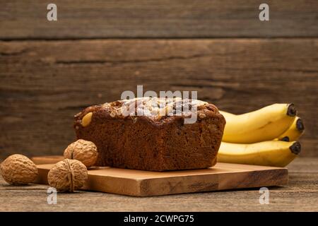 Bananenkuchen werden auf einem Holztablett auf einem Holztisch platziert, mit Bananen und Walnüssen als Hintergrund. Stockfoto