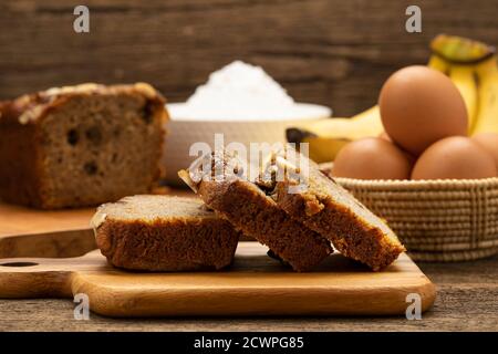 Der geschnittene Kuchen wird auf einem Holztablett auf einem Holztisch mit einer Banane, einem Ei und einem Mehl als Hintergrund platziert. ​​banana Stockfoto