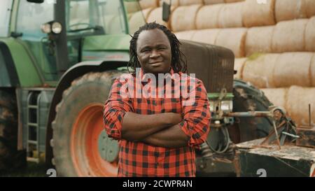 Junger afrikanischer Bauer mit gekreuzten Händen über der Brust vor Traktor und Heuhaufen. Hochwertige Fotos Stockfoto