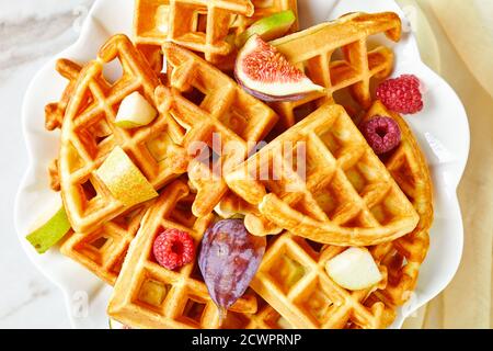 Belgische Waffeln mit frischen Beeren, Birnen, Feigen auf einem blauen Teller auf einem weißen Holztisch, flach liegend, Nahaufnahme Stockfoto