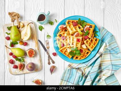 Belgische Waffeln mit frischen Beeren, Birnen, Feigen auf einem blauen Teller auf einem weißen Holztisch mit Zutaten auf einem Schneidebrett, flach liegend, horizontale Ansicht Stockfoto