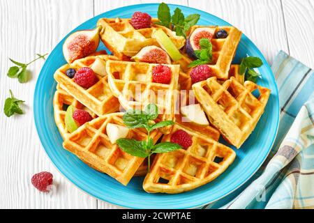 Nahaufnahme von frisch gebackenen belgischen Waffeln mit frischen Beeren, Birnen, Feigen auf einem blauen Teller auf einem weißen Holztisch Stockfoto