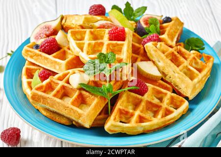 Belgische Waffeln mit frischen Beeren, Birnen, Feigen auf einem blauen Teller auf einem weißen Holztisch, Nahaufnahme Stockfoto