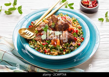 Marokkanischer Lammeintopf mit herbed Pearl Couscous und Granatapfelkernen in einer blauen Schüssel auf einem weißen Holztisch, horizontale Ansicht von oben, Nahaufnahme Stockfoto