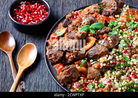 Marokkanischer Lammeintopf mit gekräuselten Perlmutt-Couscous und Granatapfelkernen auf einem schwarzen Teller auf einem dunklen Holztisch, horizontale Ansicht von oben, Nahaufnahme Stockfoto