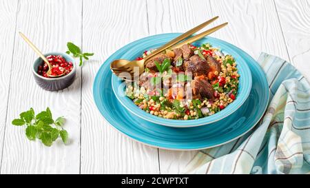 Marokkanischer Lammeintopf mit herbed Pearl Couscous und Granatapfelkernen in einer blauen Schüssel auf einem weißen Holztisch, Landschaftsansicht von oben, Nahaufnahme Stockfoto