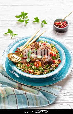 Marokkanischer Lammeintopf mit herbed Pearl Couscous und Granatapfelkernen in einer blauen Schüssel auf einem weißen Holztisch, vertikale Ansicht von oben Stockfoto