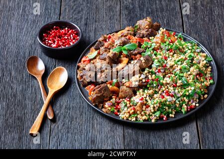 Lammeintopf mit gekräuselten Perlmutt-Couscous und Granatapfelkernen auf einem schwarzen Teller auf einem dunklen Holztisch, horizontale Ansicht von oben Stockfoto