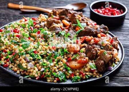 Marokkanischer Lammeintopf mit gekräuselten Perlmutt-Couscous und Granatapfelkernen auf einem schwarzen Teller auf einem dunklen Holztisch, horizontale Ansicht von oben, Nahaufnahme Stockfoto