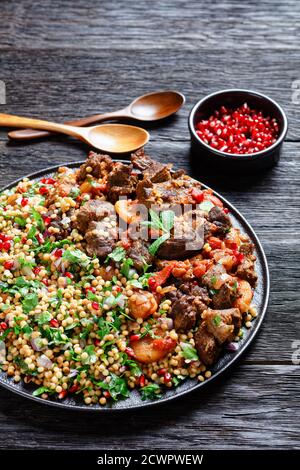 Marokkanischer Lammeintopf mit herbed Pearl Couscous und Granatapfelkernen auf einem schwarzen Teller auf einem dunklen Holztisch, vertikale Ansicht von oben Stockfoto