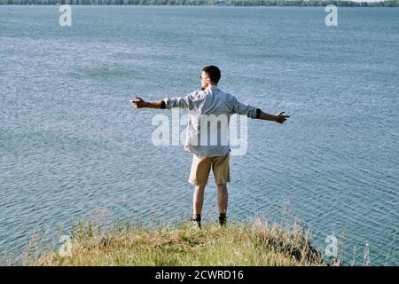 Rückansicht des jungen Mannes im Hemd stehend mit ausgestreckt Arme am Rand der Klippe und betrachten wunderschöne Wasserlandschaft Stockfoto