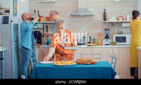 Frauen mittleren Alters und ältere Senioren haben Spaß daran, gemeinsam den Esstisch in der Küche zu stellen, während Männer im Hintergrund reden und ein Glas Weißwein während eines entspannten Familientages trinken. Stockfoto