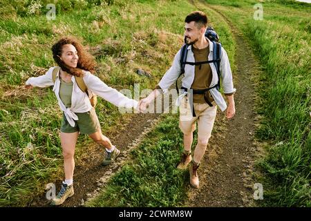 Oben Ansicht von jungen Paar Wanderer mit Rucksäcken halten Hände und gehen über Feld mit wildem Gras Stockfoto
