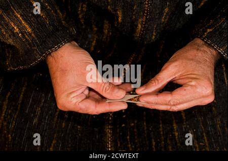 Mann mit dunkelgrüner Chilaba, stellt Zigarettenmund in die Hand, bereitet Haschisch Zigarre mit weißem Rauchpapier, Haschisch Zigarre Vorbereitung Stockfoto