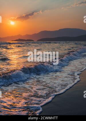 Farbenfrohe Szene von Wellen, die bei Sonnenuntergang auf den Strand von Iztuzu stürzen, mit Bergen am Horizont, die von der untergehenden Sonne umraunt werden. Stockfoto