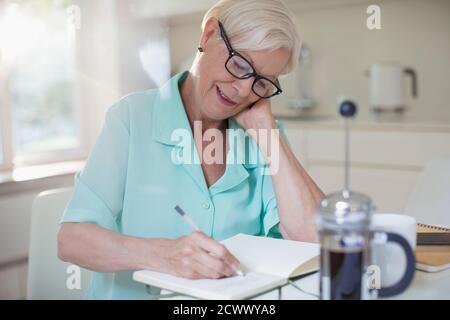 Ältere Frau, die in der morgendlichen Küche im Tagebuch schreibt Stockfoto