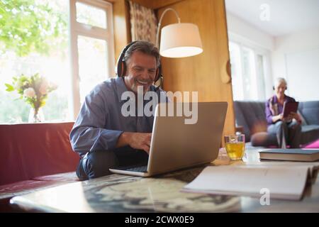Glücklicher älterer Mann mit Kopfhörern und Laptop im Wohnzimmer Stockfoto